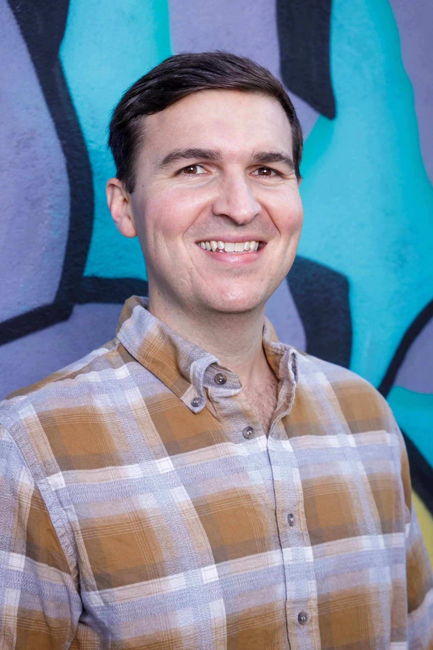 A smiling man with dark hair, wearing a plaid shirt, standing in front of a colorful graffiti wall.