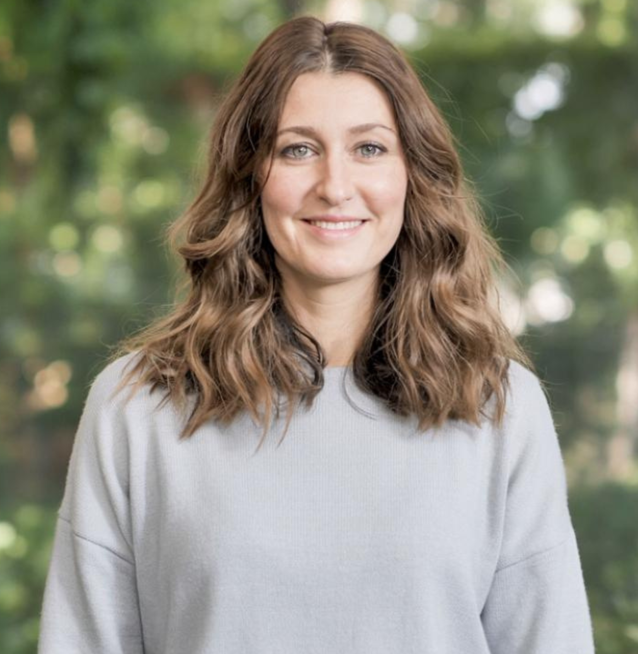 A woman with long wavy brown hair and blue eyes, smiling, outdoors with blurred trees in the background Revamp