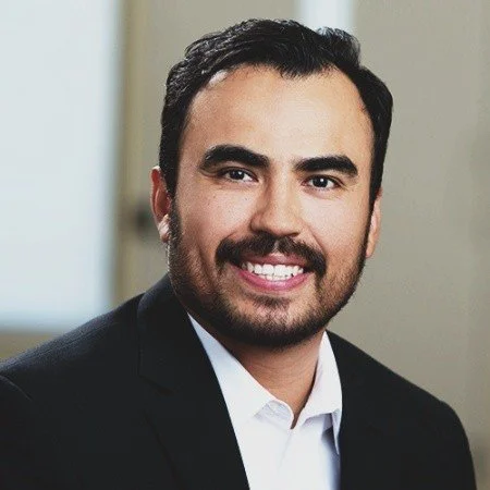 Portrait of a man with dark hair and facial hair, smiling in formal attire.