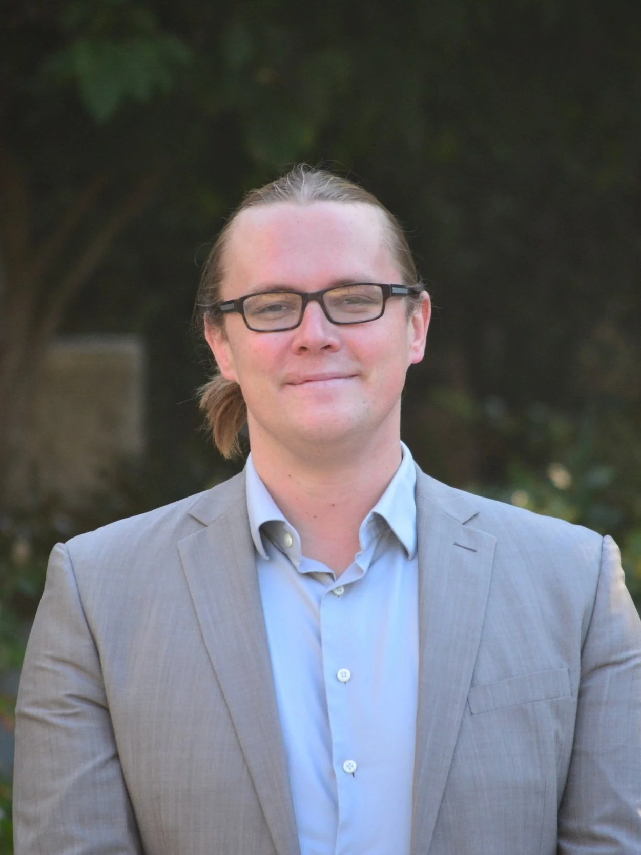 A man with glasses, wearing a light gray suit and blue shirt, standing outdoors with trees in the background.