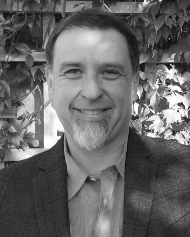 A man with short dark hair and a goatee, smiling, wearing a blazer and collared shirt, standing outdoors with leafy plants in the background.