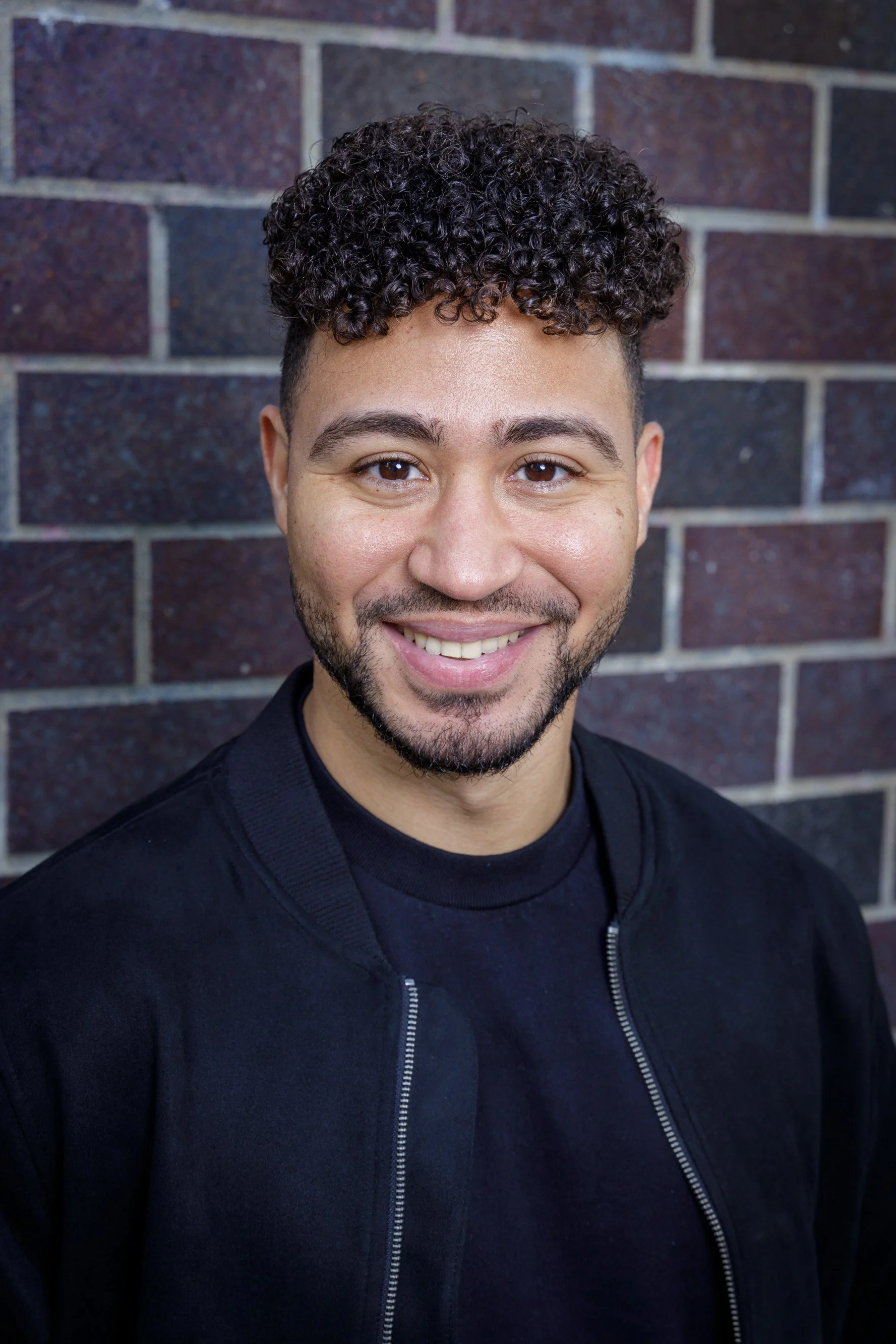 A smiling man with curly hair, beard, and wearing a black jacket, standing against a brick wall.