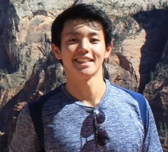 Smiling young man with black hair standing outdoors in front of a rocky canyon landscape, wearing a blue and gray shirt with sunglasses hanging from his collar.