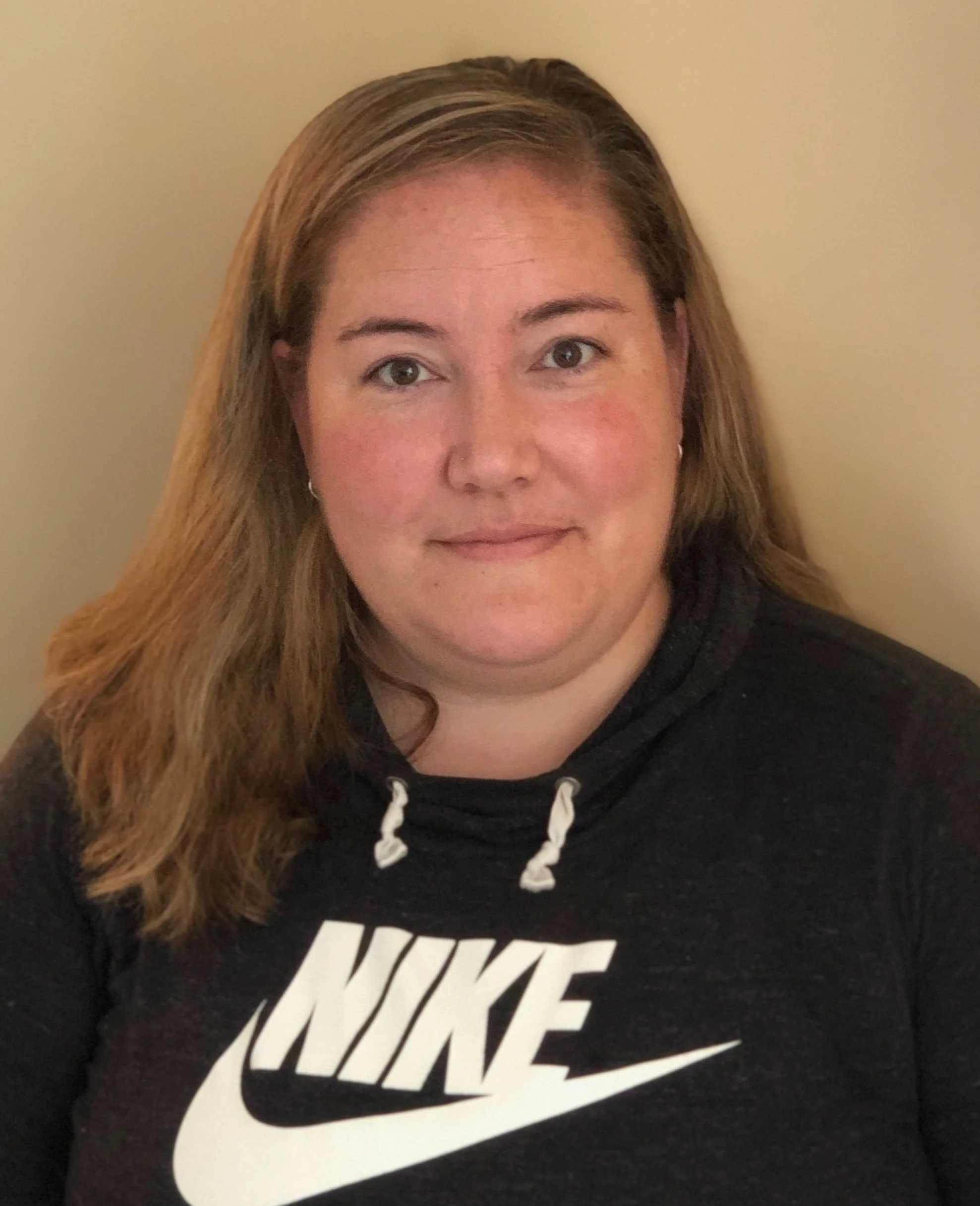 A woman with light skin and long, wavy brown hair wearing a black Nike hoodie with a white Nike logo, sitting against a beige wall.