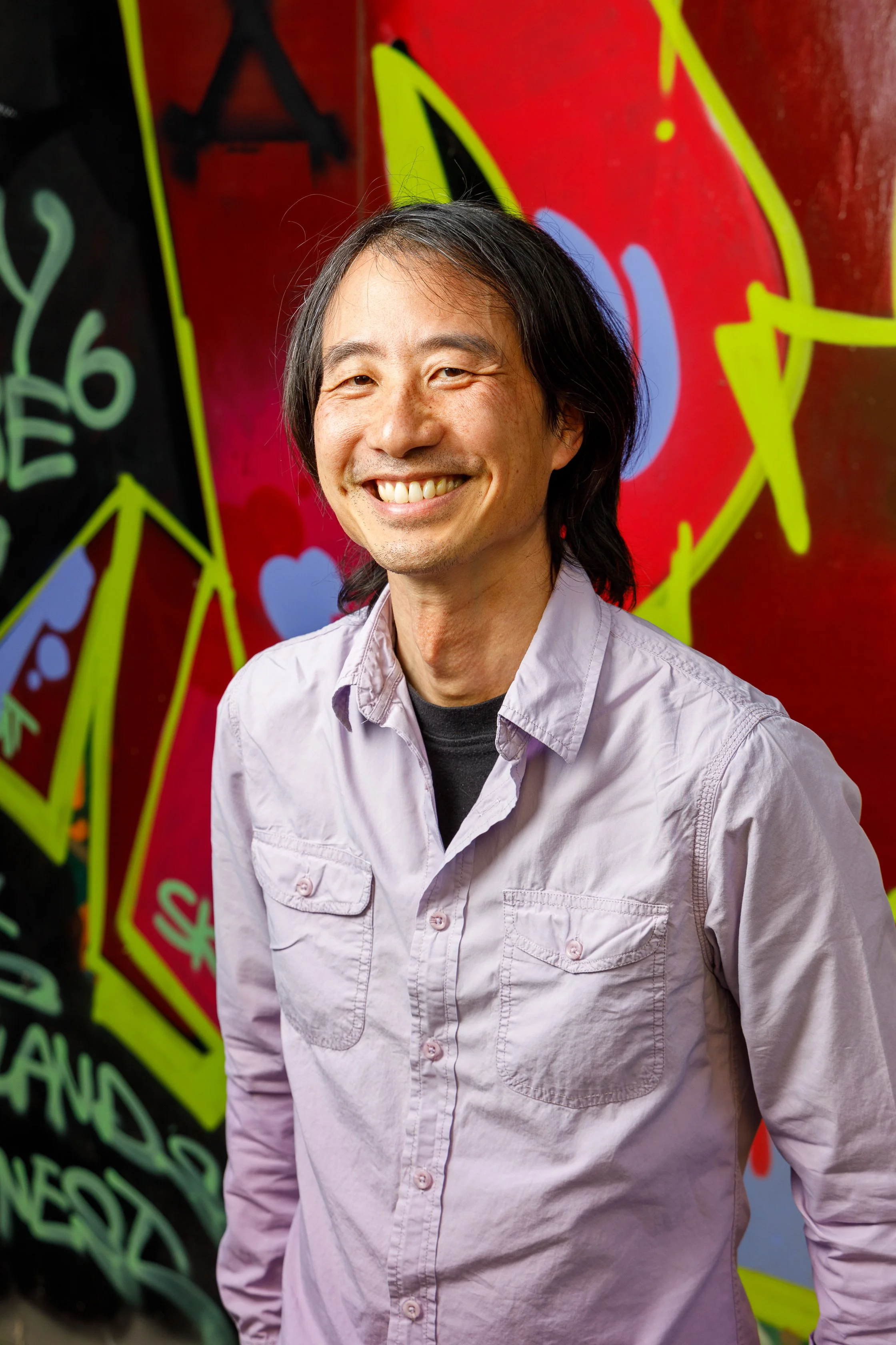 A smiling man with black hair wearing a light pink button-up shirt standing in front of a colorful graffiti wall with yellow, black, pink, purple, and green accents.