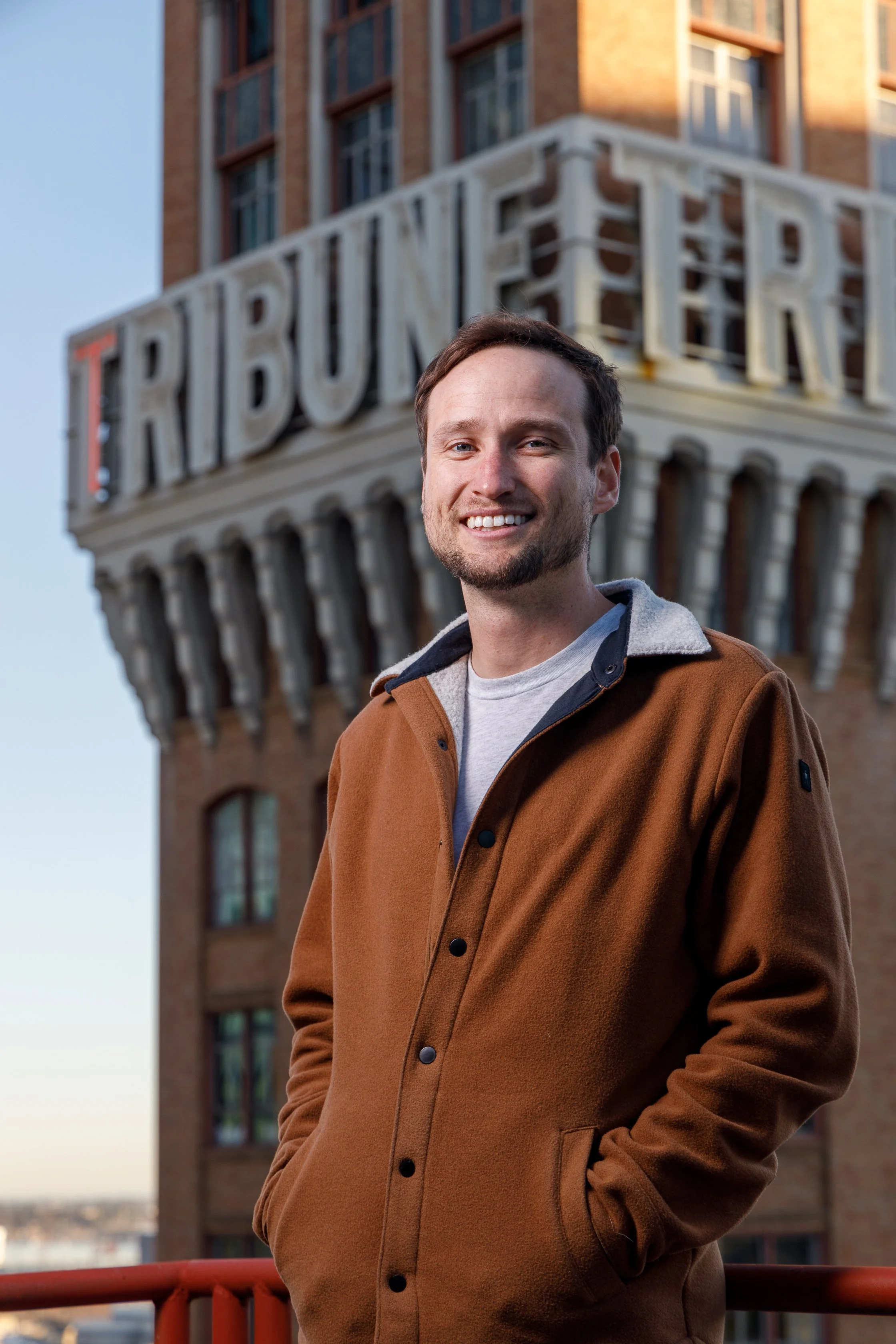 A smiling man in a brown jacket standing outside with a building and the RIBURN sign in the background.