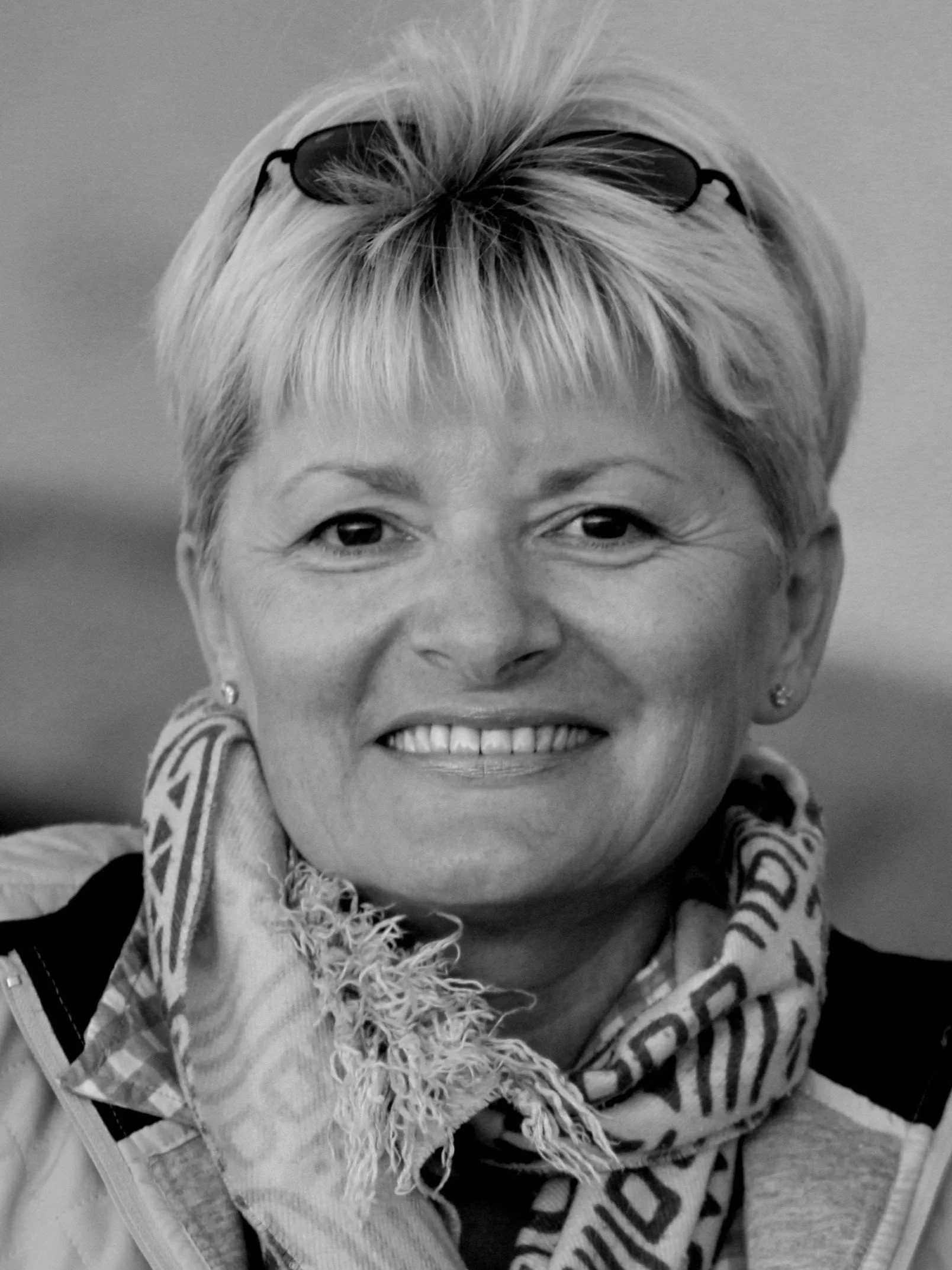 Black and white close-up photo of a smiling woman with short, light-colored hair, wearing glasses on her head, earrings, and a patterned scarf around her neck.