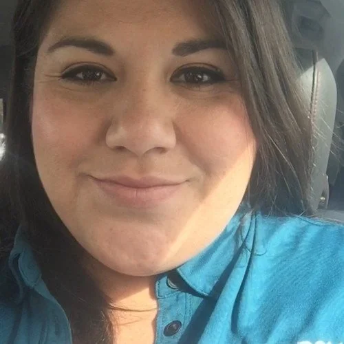 Close-up of a woman smiling, sitting inside a car, wearing a blue shirt.