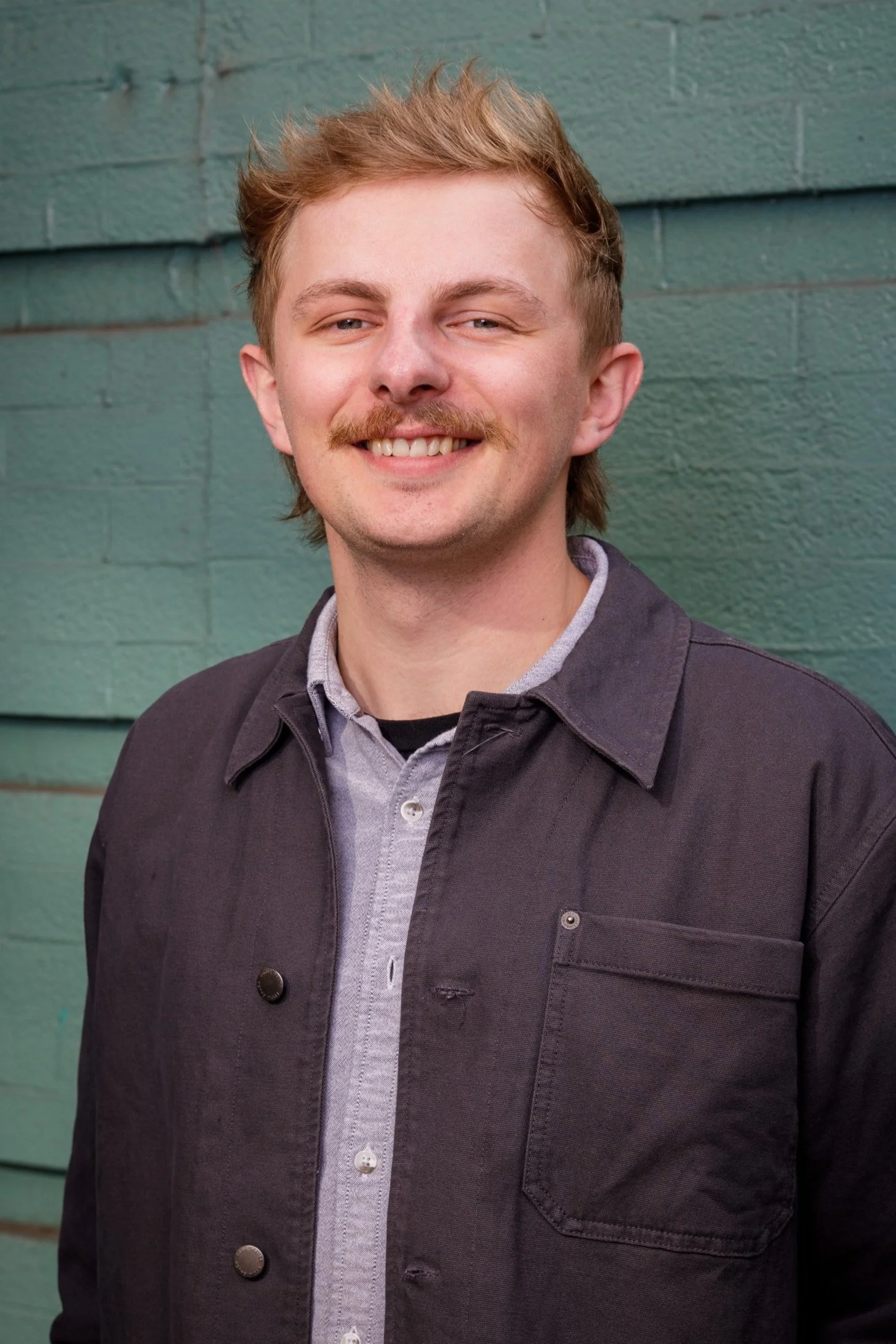 A young man with light skin, blond hair, and a mustache, smiling, standing against a green textured wall, wearing a gray shirt and a black jacket.