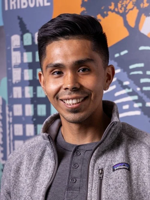 Young man smiling, wearing a gray Patagonia fleece jacket, standing in front of a colorful cityscape mural.