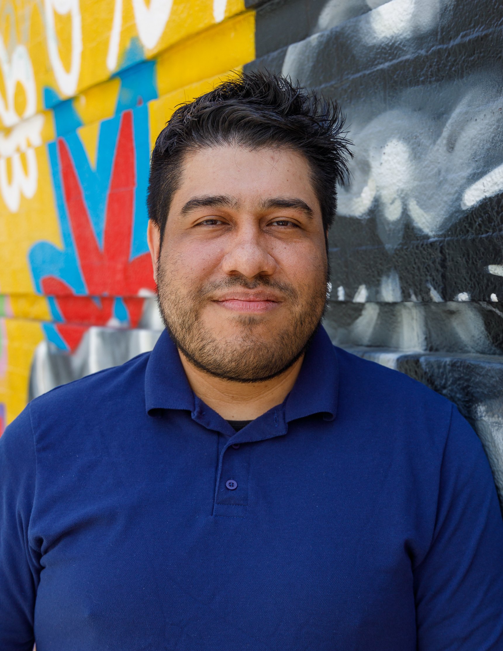 A man with dark hair and a beard wearing a navy blue polo shirt, standing in front of a colorful mural on a brick wall.