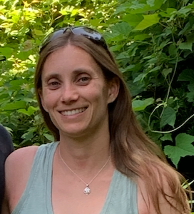 A smiling woman with long brown hair wearing sunglasses and a light blue sleeveless top, standing outdoors among green foliage.
