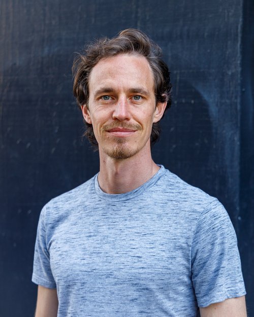 Portrait of a man with curly brown hair, light beard, wearing a gray t-shirt, standing outdoors against a dark background.