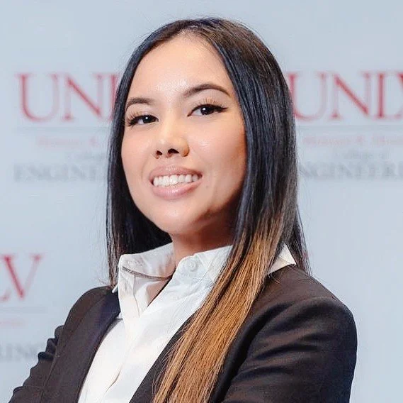 A young woman with long dark hair, smiling and sticking her tongue out slightly, wearing a white shirt and dark blazer, standing in front of a blurred background with text.