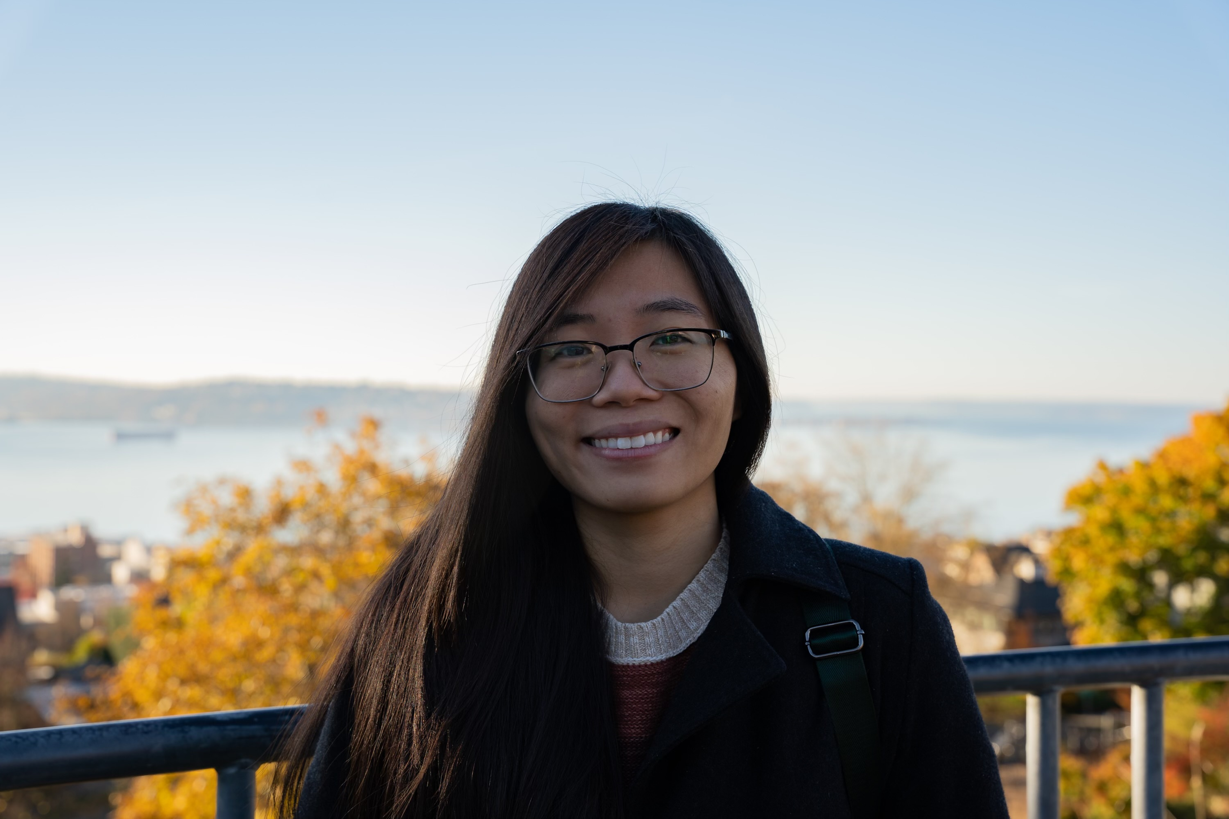 A young woman with long dark hair, wearing glasses and a black coat, smiling outdoors with autumn trees and a body of water in the background on a clear day.