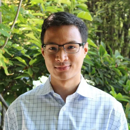 A young man wearing glasses and a light blue checkered shirt standing outdoors in front of green leafy plants.