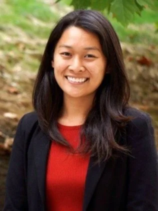 A woman with long dark hair smiling outdoors, wearing a black blazer over a red top, with green foliage in the background.
