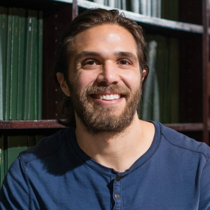A smiling man with a beard and long hair, wearing a blue shirt, standing in front of bookshelves filled with green books.