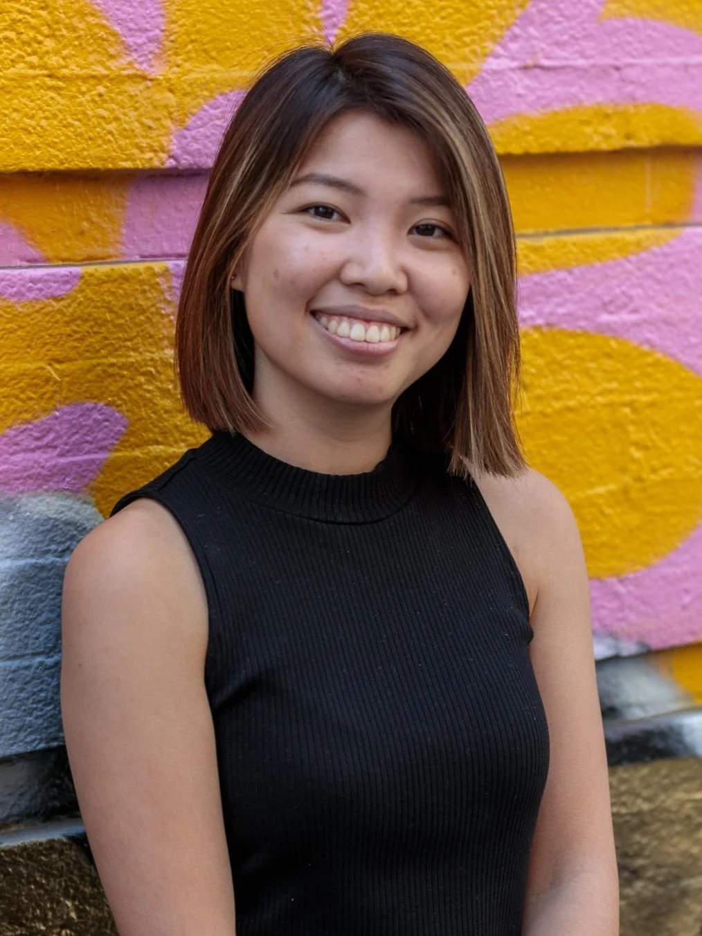 A young woman smiling in front of a colorful graffiti wall with pink, yellow, and purple paint.