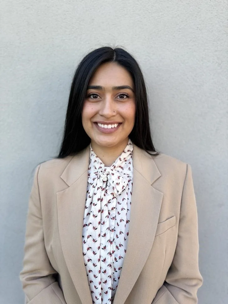 A woman with long dark hair in a beige blazer and patterned blouse, smiling in front of a plain wall.