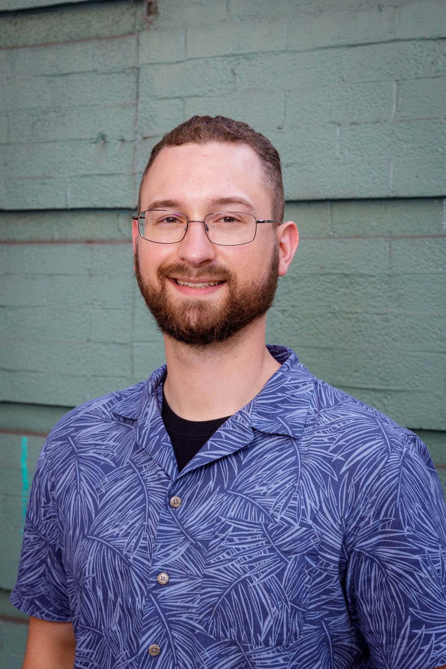 Portrait of a man with glasses and a beard, smiling, wearing a blue patterned shirt, standing in front of a green painted brick wall.