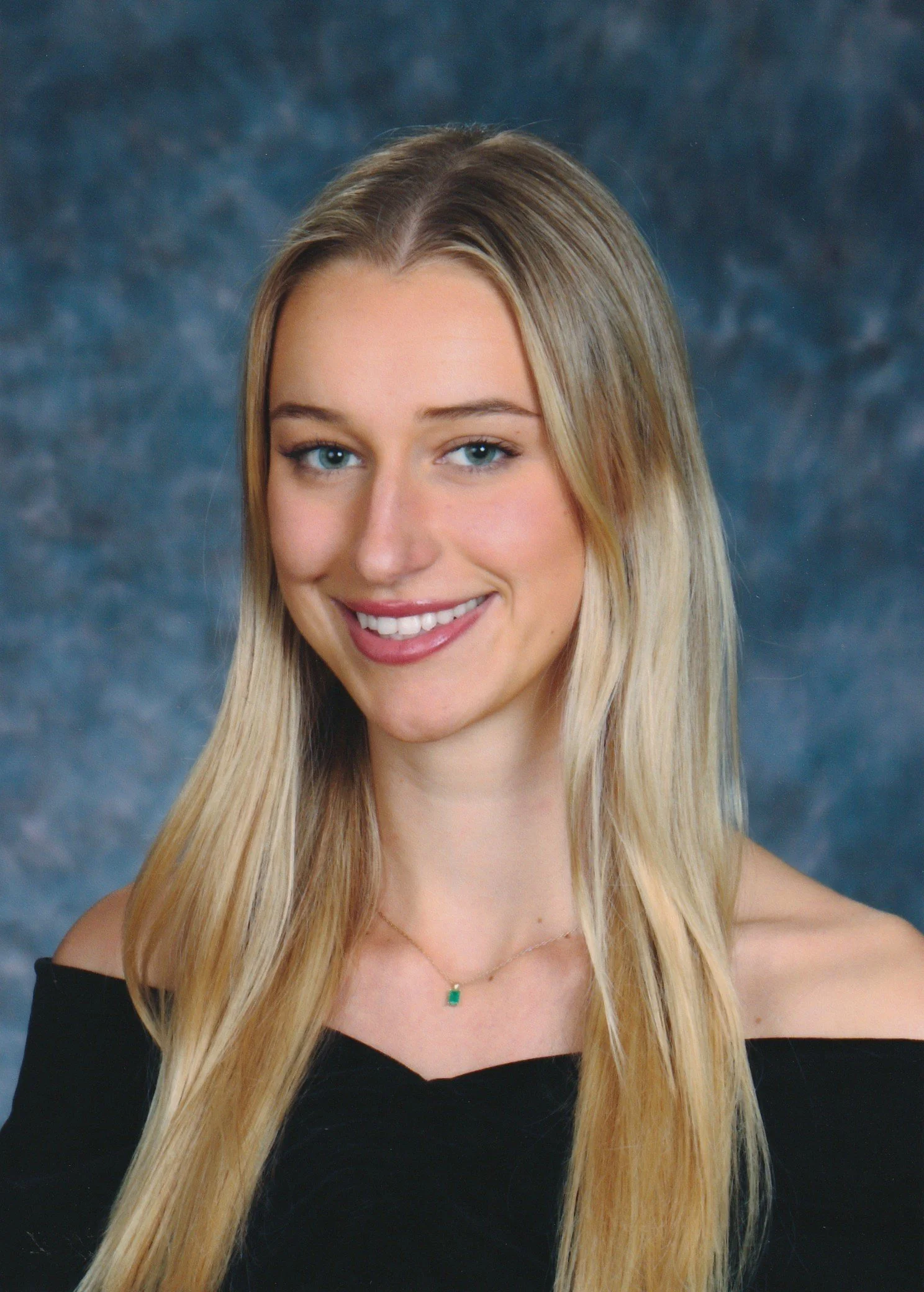 A young woman with long blonde hair, blue eyes, and a warm smile wearing an off-the-shoulder black top and a delicate necklace with a small green pendant, posed against a blue mottled background.