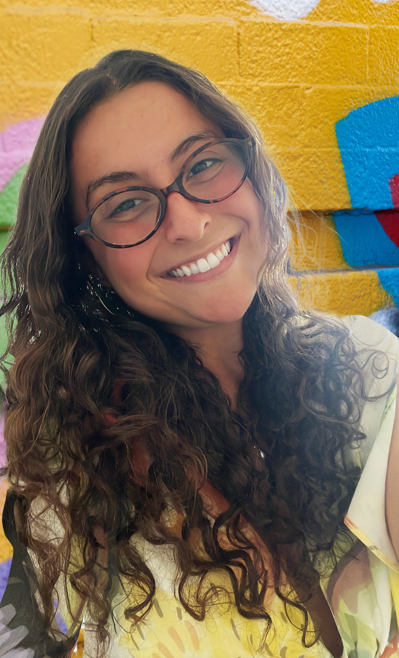 A young woman with curly brown hair wearing glasses smiling in front of a colorful graffiti wall.
