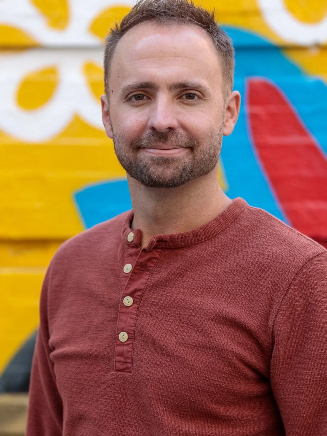 A man with short brown hair, trimmed beard, wearing a red long-sleeve shirt with buttons, standing outdoors in front of a colorful mural with yellow, white, blue, and red designs.