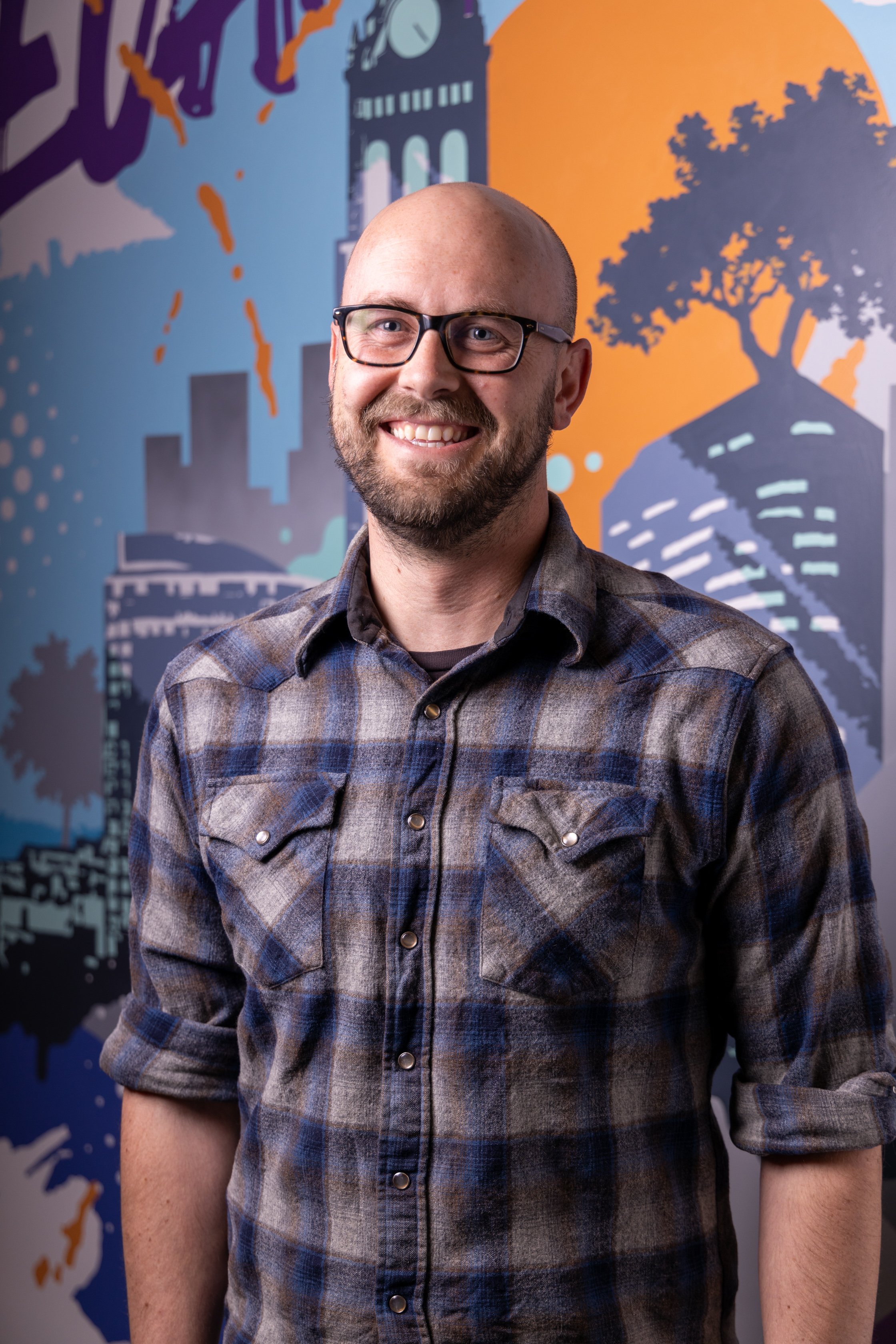A smiling man with glasses and a beard, wearing a plaid shirt, standing in front of a colorful mural featuring silhouette city buildings, trees, a clock tower, and abstract shapes with a sunset background.