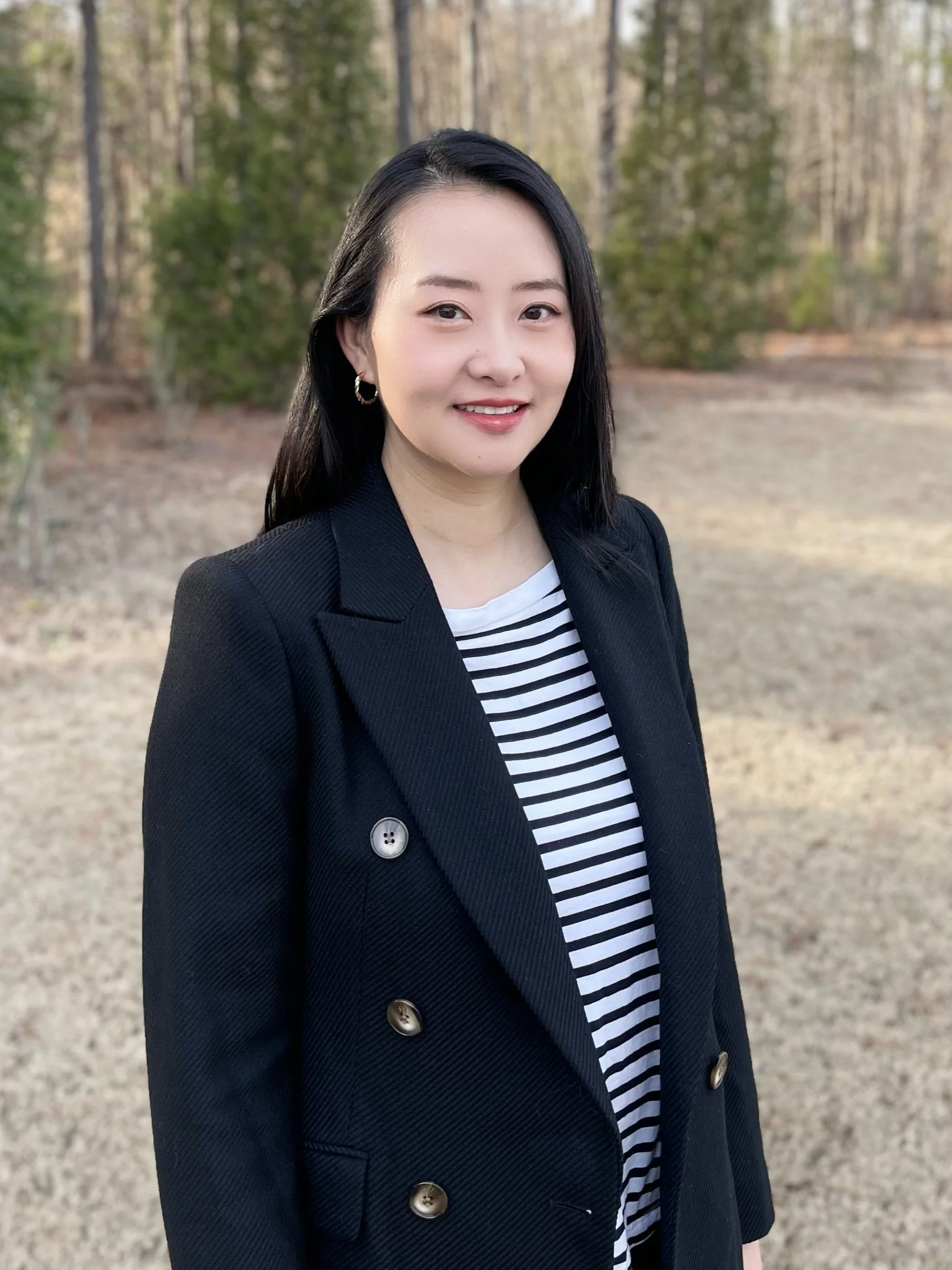 A woman with black hair smiling outdoors, wearing a black blazer, striped shirt, and hoop earrings, with a forested background.