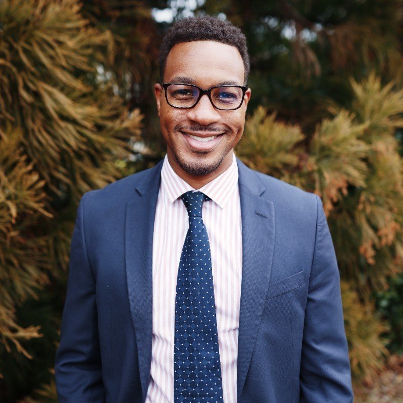 A smiling man with glasses wearing a navy suit, white striped shirt, and a dotted tie, standing outdoors with autumn-colored trees in the background.