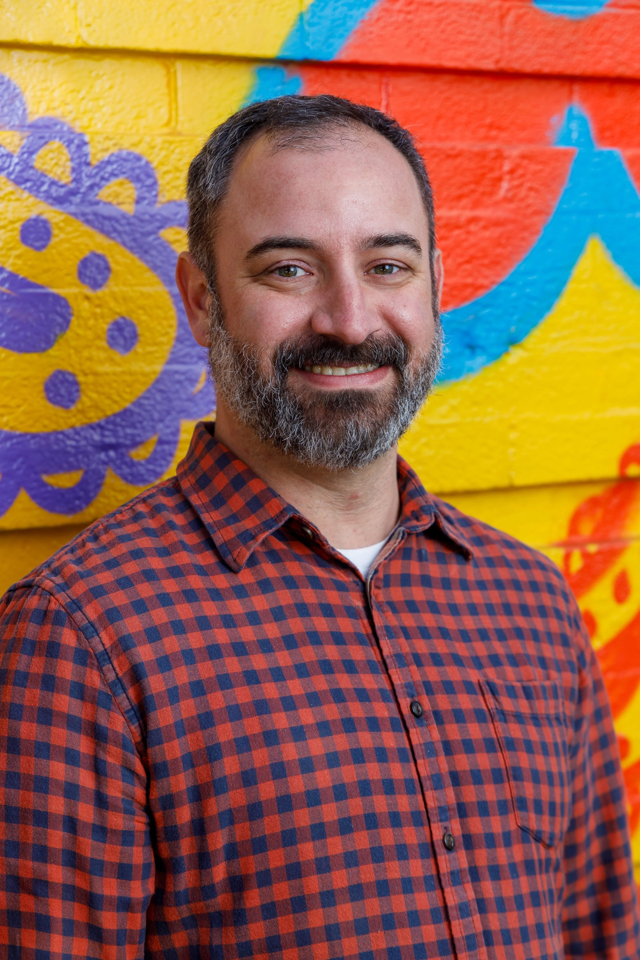 A smiling man with a beard and short hair standing in front of a colorful, painted brick wall.