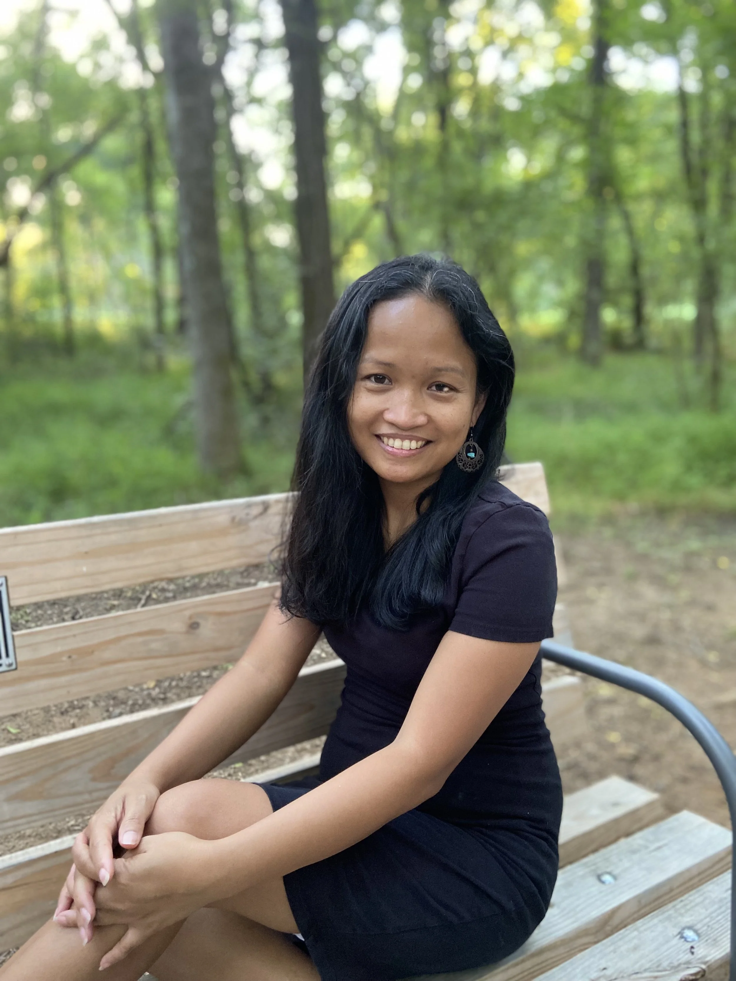 A woman with long black hair smiling, sitting on a wooden bench in a wooded park, wearing a black dress and earrings.