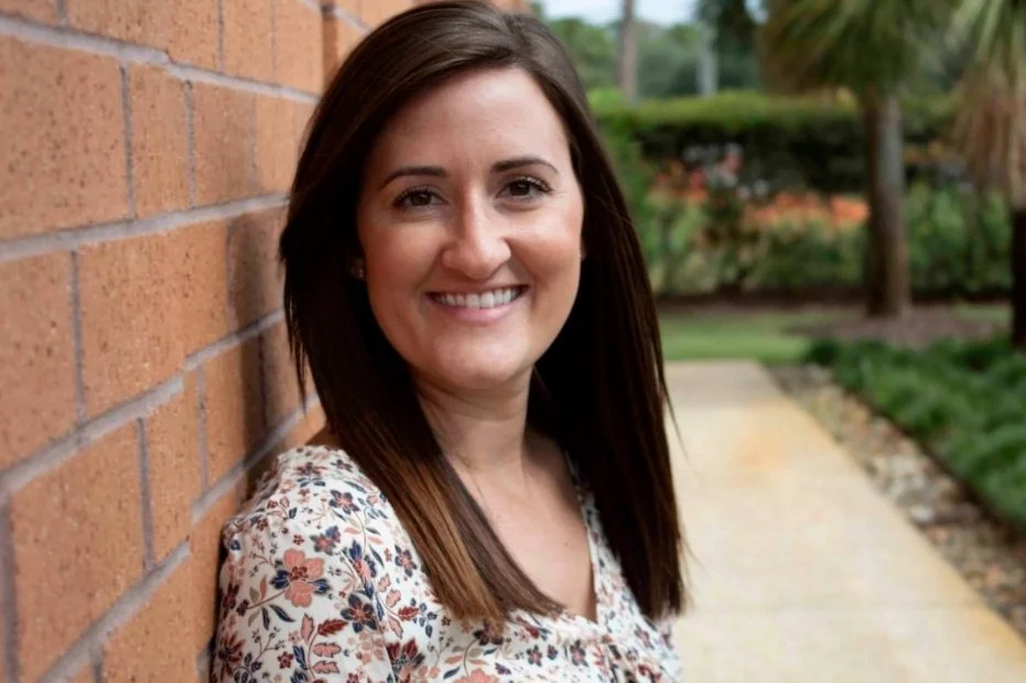 A woman with shoulder-length brown hair, smiling, wearing a floral shirt, leaning against a brick wall outdoors with greenery and trees in the background.