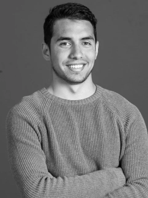 Black and white portrait of a young man with short dark hair, a light beard, wearing a knit sweater, smiling with arms crossed against a plain background.