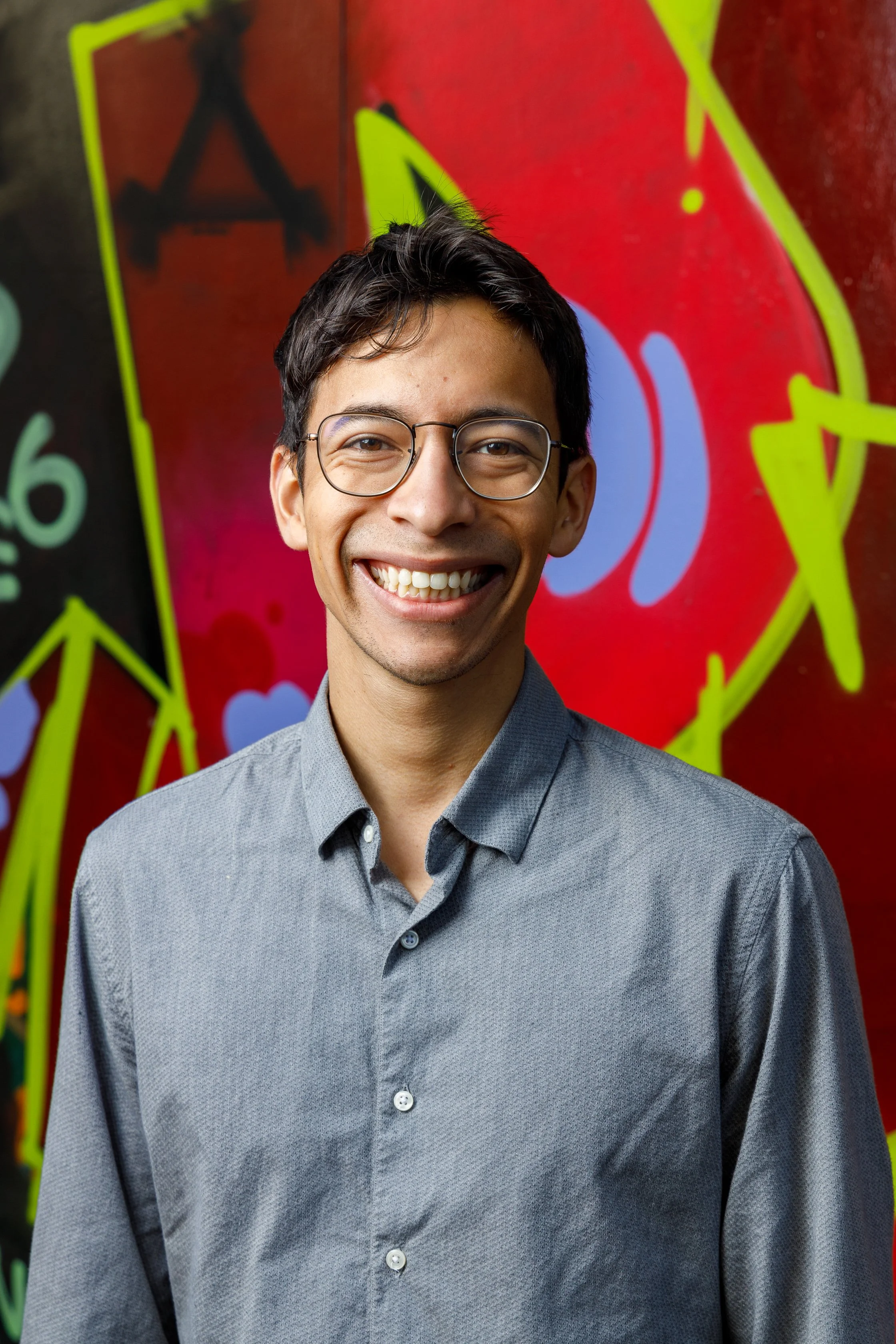 A smiling man with glasses wearing a gray shirt standing against a colorful graffiti wall.