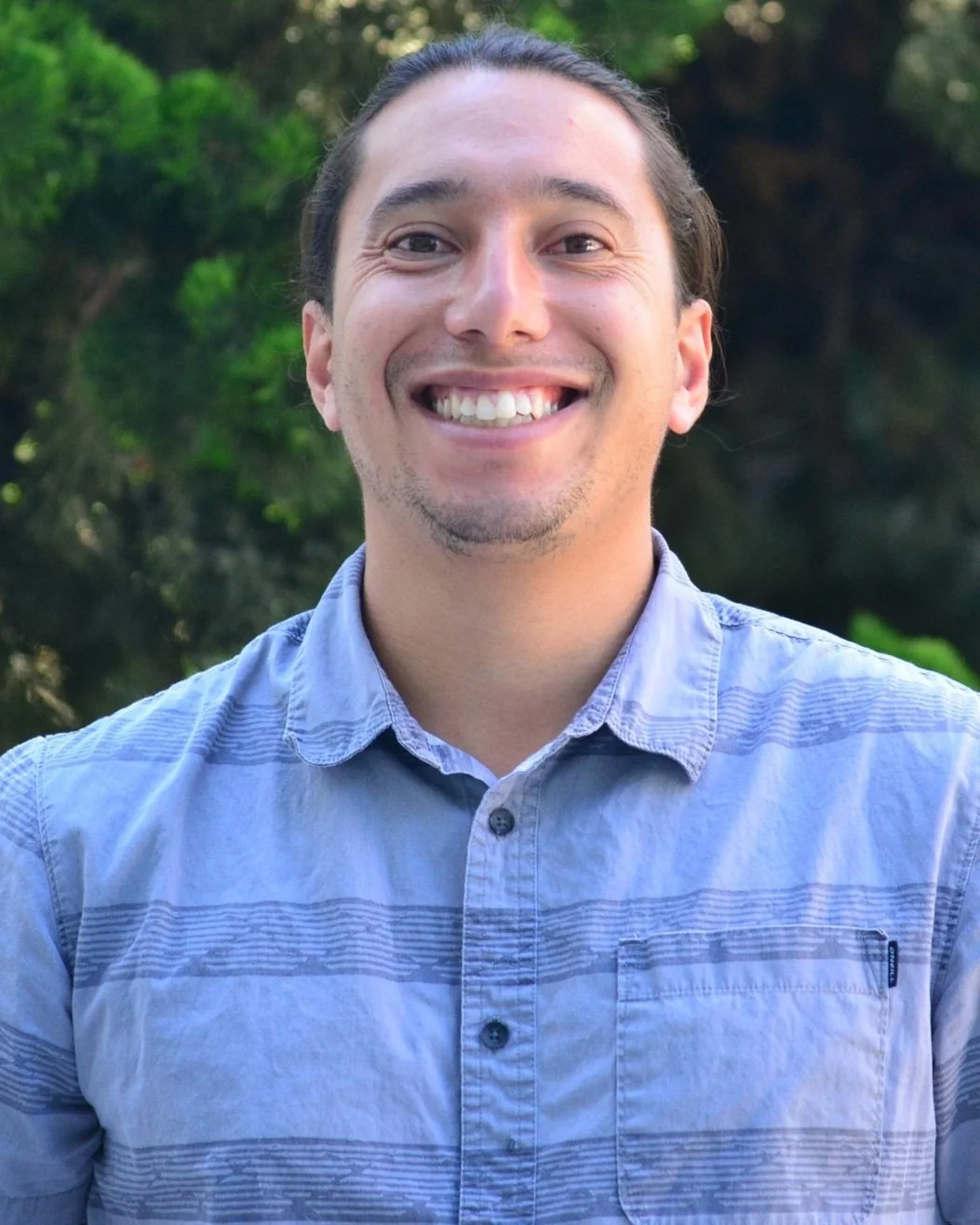 A smiling man with long dark hair tied back, wearing a light blue button-up shirt, standing outdoors with green trees in the background.