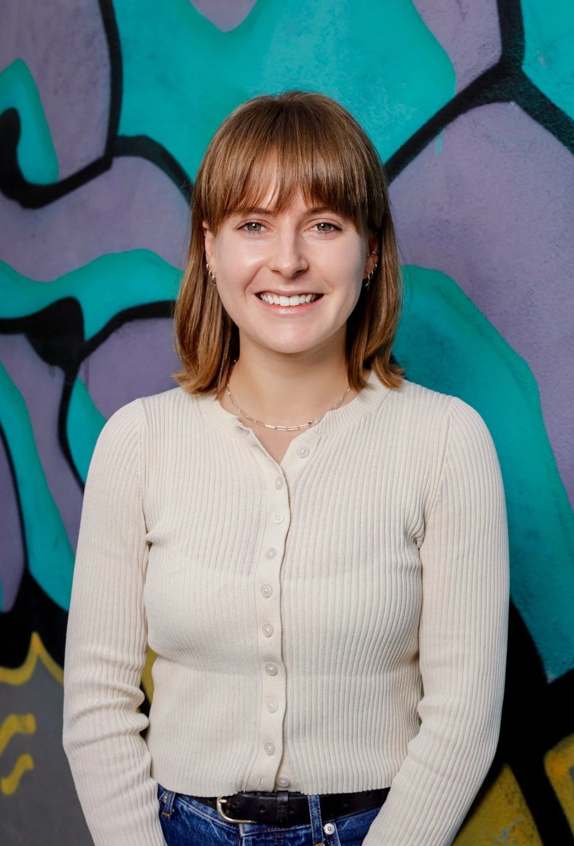 A young woman with shoulder-length brown hair, wearing a cream-colored button-up ribbed sweater and blue jeans, smiling in front of a colorful abstract graffiti wall.