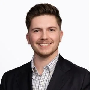 A young man with short brown hair, wearing a dark blazer and a checkered shirt, smiling against a plain white background.