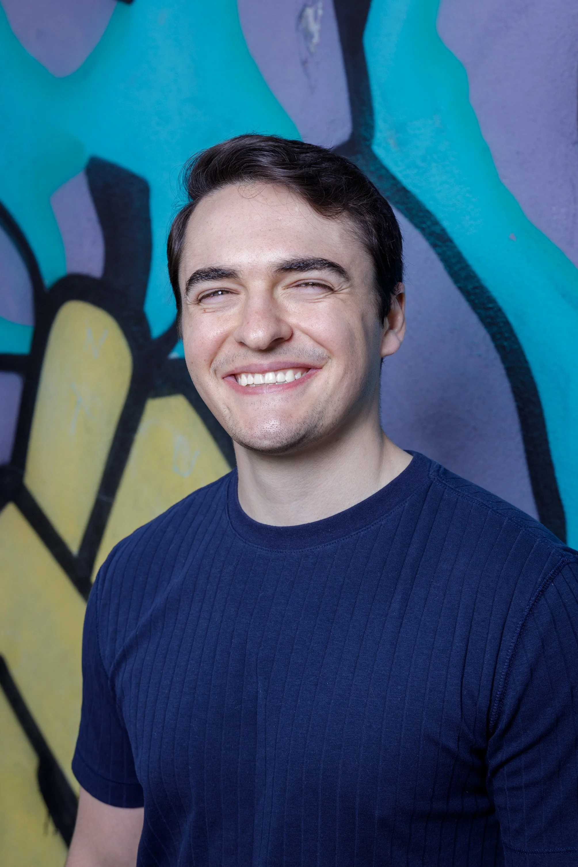 Smiling young man with dark hair standing in front of a colorful graffiti wall.