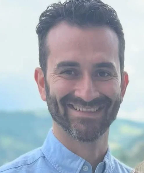 Smiling man with dark hair and beard wearing a blue collared shirt, outdoor background.