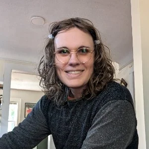 A woman with curly hair and glasses smiling indoors.