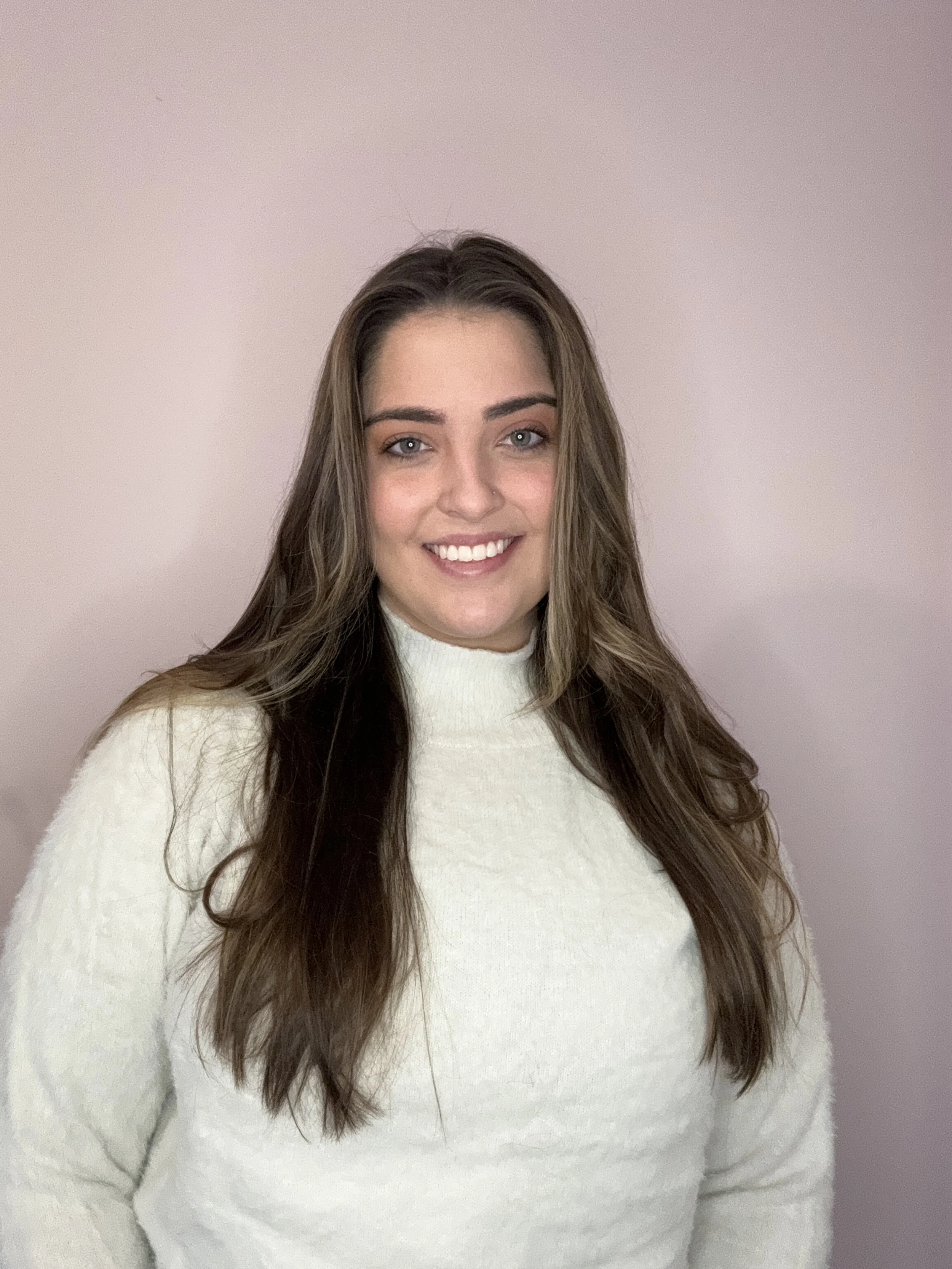A woman with long brown hair wearing a cream-colored sweater, smiling at the camera, standing against a plain pink background.