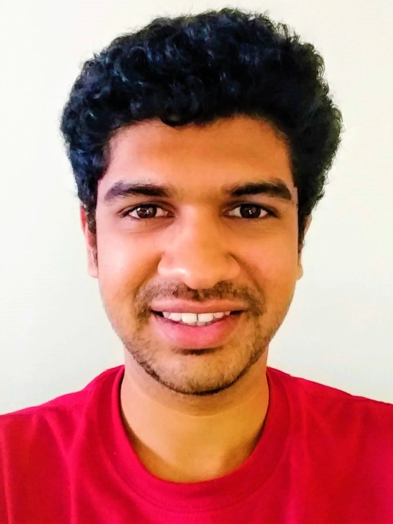 A smiling young man with curly black hair, wearing a red shirt, standing against a plain light-colored background.