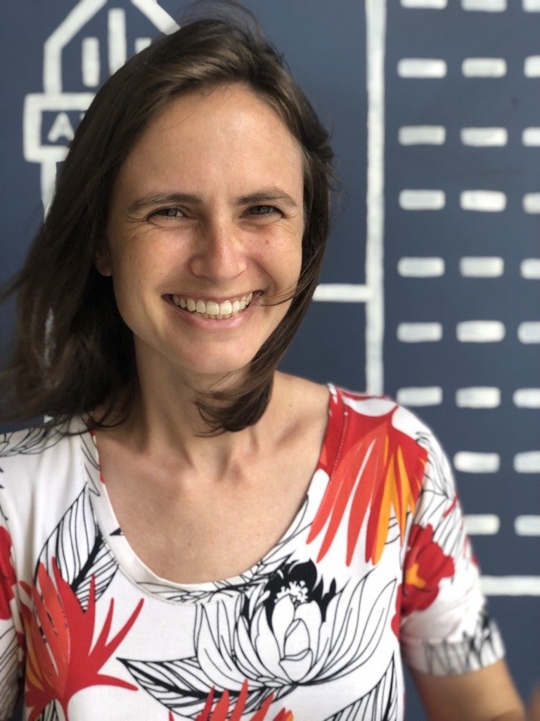 A woman smiling in front of a blue wall with white abstract drawings. She has shoulder-length brown hair and is wearing a white shirt with a red, black, and yellow floral pattern.