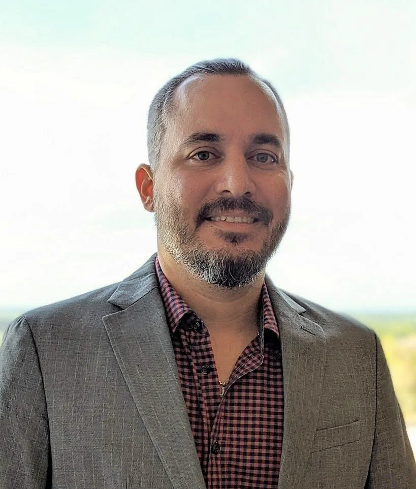 A man with short gray hair, beard, and mustache smiling, wearing a gray blazer and a red checkered shirt, standing in front of a bright background with a faint landscape view.