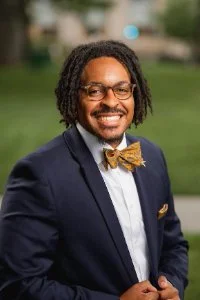 A smiling man with glasses and dreadlocks wearing a navy blazer, white shirt, and gold bow tie, outdoors with greenery in the background.