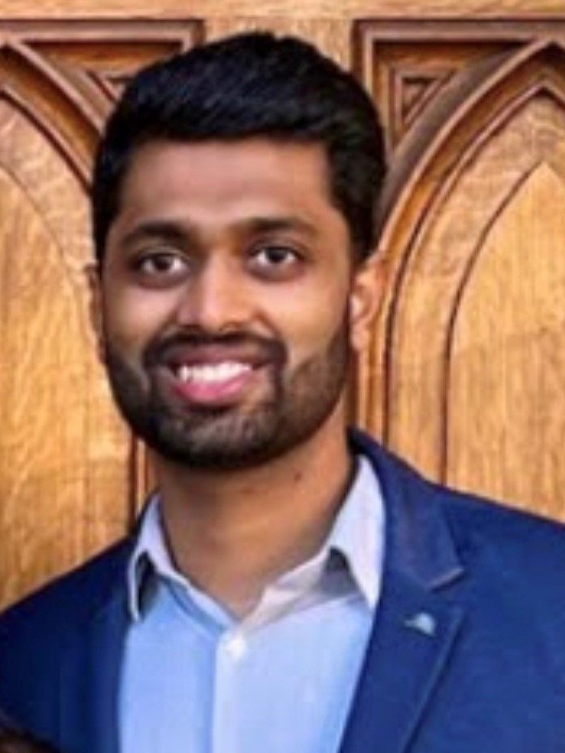 A man with dark hair and a beard, wearing a blue suit jacket and a light-colored shirt, smiling in front of a wooden background.