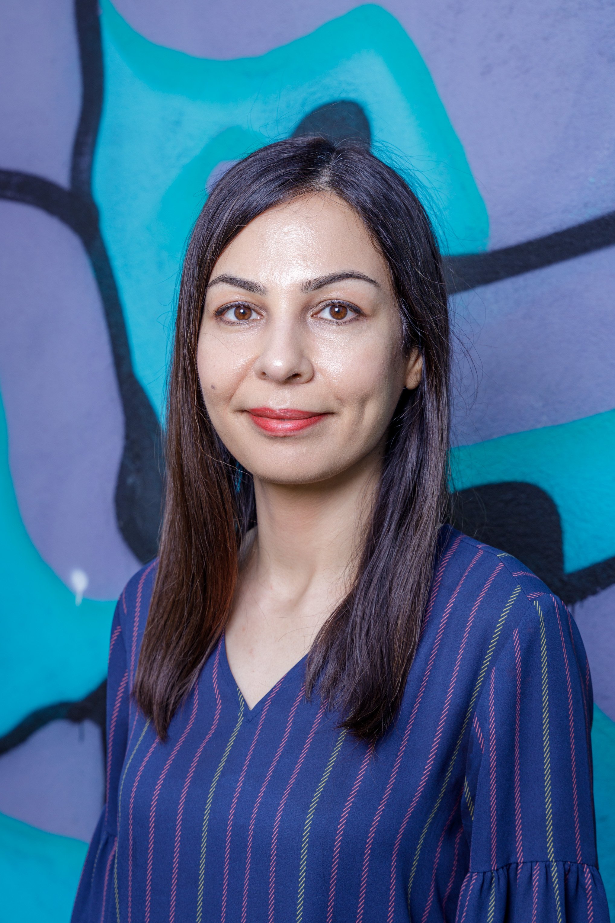 A woman with long dark hair and light skin standing in front of a colorful graffiti wall, wearing a dark blue striped blouse and smiling at the camera.