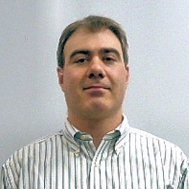A man with short brown hair, wearing a white striped button-up shirt, standing against a plain light-colored wall.